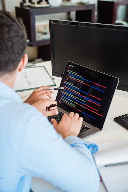 A close-up view of a person's hands operating a laptop with a sleek, modern design, positioned on a dark table. The laptop's screen displays lines of colorful code and text on a dark background, indic