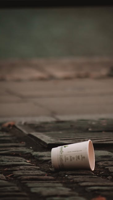 A large pile of mixed household waste and rubbish is gathered beside a metal railing on a paved urban street, with various discarded items including cardboard boxes, plastic bags, paper, and packaging